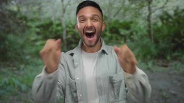 Hispanik adam şaşkınlığını ifade ediyor, ağzı ve elleri açık, yeşil bir park ortamında, doğal ortamda gençlik neşesi ve doğallığı kapsıyor..