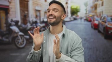 Kentsel bir sokak ortamında enerjik bir şekilde gülümseyen ve el kol hareketi yapan genç İspanyol adam, neşeli bir şehir ortamında mutluluk ve pozitiflik taşıyor..