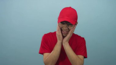 Kırmızı gömlekli ve şapkalı, gözlüklü, mavi stüdyoda elleri yanaklarında, diş ağrısı çeken genç bir adam..