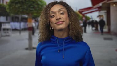 Mavi kapüşonlu, kıvırcık saçlı bir kadın şehir sokaklarında bir objeye iki eliyle işaret ediyor..