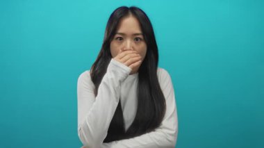 Genç Çinli kadın, izole edilmiş mavi arka plan duvarına karşı ağzı şaşkınlıkla kapatıyor. Uzun siyah saçlı ve beyaz giysili bir kadın..