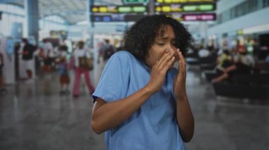 Mavi önlüklü kadın endişeli bakışlarla havaalanı terminalinin çıkış panosuna el sıkıştı; endişe.