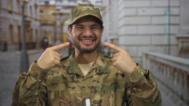 Kamuflaj üniformalı bir adam, şehir sokaklarında güler yüzlü ve yanakları işaret eder..