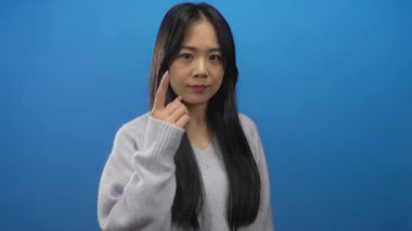 Hırkalı genç bir kadın mavi arka plan duvarının üzerinde parmak göze işaret ediyor..