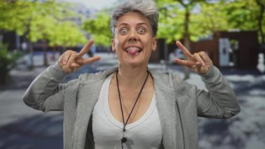 Kısa saçlı, gri kapüşonlu bir kadın gülümsüyor ve ağaçlarla kaplı bir sokakta olumlu bir hareket sergiliyor; iyimserlik.