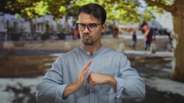 Latin kökenli genç adam ağaçların altında yumruğunu sıkıyor. Şehir ortamında mavi gömlekli ve gözlüklü.