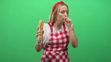 Kırmızı kareli önlük giyen kadın şef sol elinde bir baget tutuyor ve yeşil ekranlı bir stüdyoda sağ yumruğuna öksürüyor..
