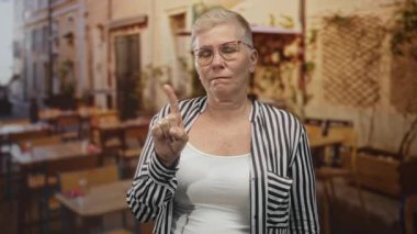 Çizgili tişörtlü ve gözlüklü bir kadın dışarıdaki kalabalık bir caddedeki restoran terasından işaret parmağıyla işaret ediyor..