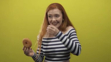 Kızıl saçlı kadın bir elinde donut, diğer elinde de çizgili bluz giyerken ağzını stüdyoda kapatıyor..