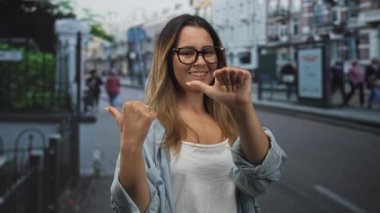 Şehir caddesinde başparmağıyla göğsünü gösteren, gözlük takan ve beyaz kolsuz bluz giyen, kendine güvenen bir kadın..