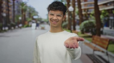 Beyaz kazaklı ve gözlüklü bir adam, güneşli şehir caddesinde uzanan avuç içi sunar; sıcak karşılama.