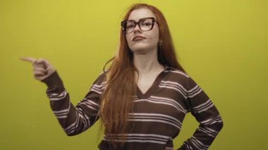 Stüdyoda eli kalçasında, büyük gözlüklü ve kahverengi çizgili süveter giyen, sol elini işaret eden kızıl saçlı kadın, şüphecilik yargısı.