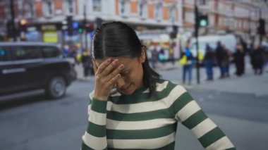 Renkli binalar ve hareketli kalabalıkla caddede dikilen kadın şehir ortamında duygularını gösteriyor ve çizgili kıyafetler giymiş genç bir bireye odaklanıyor..