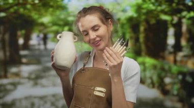 Önlüklü genç bir kadın elinde seramik bir kavanoz ve boya fırçaları tutarken, yaratıcı sürecin tadını çıkarırken, açık havada sanatsal tutku sergiliyor..