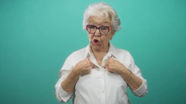 Turkuaz stüdyoda el ele tutuşan yaşlı kadın, gözlük takıyor ve yüzüklü beyaz gömlek giyiyor; sevgi dolu aile hafızası.