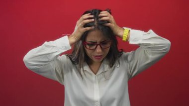 Gerginliğini ifade eden bir kadın, elleri başının üzerinde canlı kırmızı bir arkaplan önünde, beyaz gömlek ve kırmızı gözlük takıyor, duygularını canlı bir şekilde gösteriyor ve endişe ve hayal kırıklığını dile getiriyor.