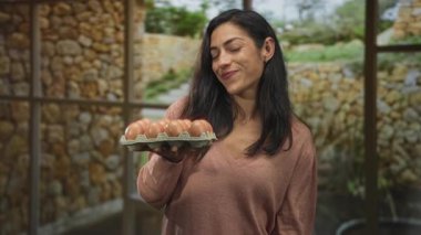 Stüdyoda pencere kenarına uzanmış taş duvarın yanında gülümseyen ve yumurta kutusunu tutan İspanyol kadın, basit bir ev sıcaklığı..