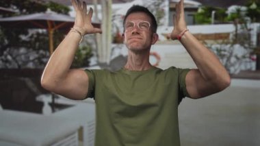 Zeytin yeşili gözlüklü bir adam havuz alanında iki elini kaldırıyor ve görünür can simidi ve şemsiyeleri var..