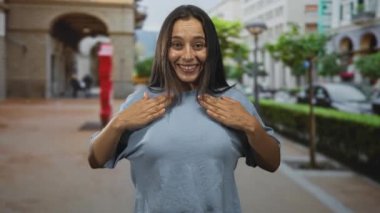 Kadın göbeğini göstermek için gömleğini kaldırırken gülümsüyor ve kemerli bina ve park halindeki arabayla iki kolunu kaldırıyor..