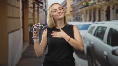 Kadın, elinde optometri deneme çerçevesi, göğsünde el, kolsuz siyah bluzlu sarışın, park halindeki araba ve binalarla dolu bir caddede duruyor, minnettarlık..