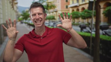 Bir şehir caddesinde ellerini açık gösteren ve geniş bir gülümseme, avuçlar görünür ve parmaklar kırmızı polo gömlek giyerken oyuncu bir pençe hareketi yayılmış;.