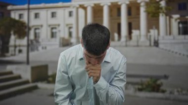 Sütunlu binanın önünde öksürük için eliyle ağzını kapatan İspanyol genç adam, sağlık endişesi.
