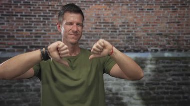 Adam iki eliyle tuğla bina duvarına karşı baş parmağını kaldırıyor, kameraya bakıyor, gözleri kapalı ve hafifçe sırıtıyor; eleştiri.