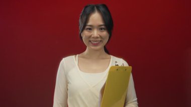 Elinde dosya olan genç Çinli bir kadın canlı kırmızı bir arka plana karşı hoş geldin jesti yapıyor. Yüzü çekici ve dost canlısı..