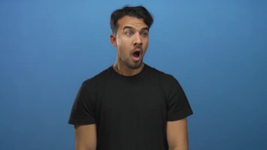 Siyah gömlek giyen İspanyol genç adam, izole edilmiş mavi arka plana karşı şaşkınlığını dile getiriyor.