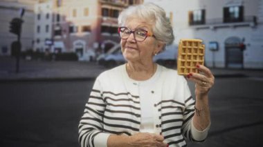 Son sınıf öğrencisi kadın, çizgili hırka ve renkli gözlük takmış, gülümserken sokakta waffle yapıyor..