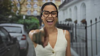 Sokak ortasında gülen İspanyol bir kadın, gözlükleriyle gülümsüyor ve kameraya neşeli bir şekilde işaret ederken üstündekini giyiyor..
