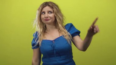Kireç yeşili arka planda, mavi elbiseli ve dağınık sarı saçlı bir kadın stüdyoda yukarı doğru işaret ediyor..