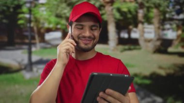 Kırmızı şapkalı adam akıllı telefonu kulağına tutarken gülümsüyor ve sokakta tablet okuyor; bağlantı.