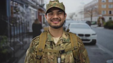 Kentsel sokak ortamında kamuflaj üniformalı, sırt çantalı ve künyeli bir adam gülümsüyor..