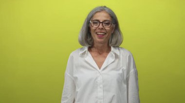 Orta yaşlı, gri saçlı, elleri sımsıkı gülümseyen, limon yeşili arka planda beyaz gömlek giyen, sevinç sıcaklığı olan kadın..