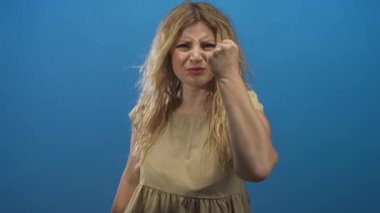 Parlak mavi stüdyodaki kameraya yumruk sallayan genç sarışın kadın, öfke meydan okuması..