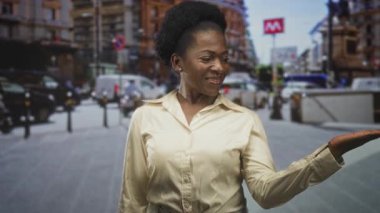 Bir metro girişinin yakınındaki işlek bir caddede el hareketleriyle avuç içi gösteren kadın, kameraya gülümseyen ve el kol hareketi yapan;.