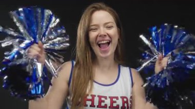 Genç beyaz amigo kız, stüdyoda siyah arka planda metalik mavi ponponları enerjik bir şekilde sallıyor..