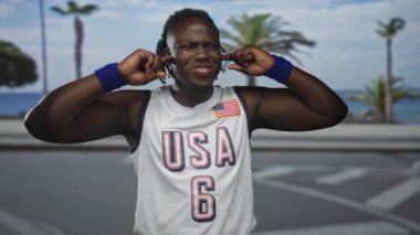 Deniz kenarındaki sokakta kulaklarını tıkayan genç bir adam Amerikan basketbol forması ve bileklikleri takıyor..