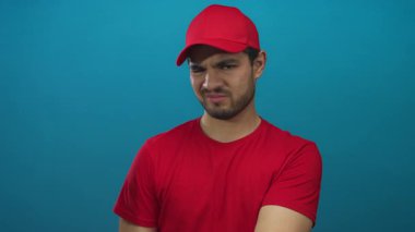 Kırmızı şapkalı ve gömlekli genç İspanyol kurye mavi stüdyoda kaşlarını çatıyor ve parmaklarını parmakla gösteriyor..