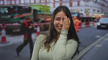 Bir şehir caddesinde, arkasında trafik konileri olan inşaat araçlarının önünde eli ile gözlerini kapayan genç bir kadın; çekingenlik.