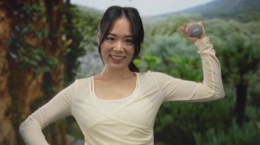 Yeşil bir parkta dambılla egzersiz yaparken gülümseyen genç Çinli kadın, özgüvenle arka planda canlı bir doğayla baş parmak hareketini gösteriyor..
