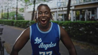 Adam mavi basketbol forması içinde beyaz bilekliklerle el çırpıyor, çalıların ve bina cephesinin olduğu bir caddede geniş bir gülümseme sergiliyor; sevinç kutlaması..