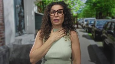 Park halindeki araçların yanında sandık başında duran ve yeşil ağaçların altında tuğla bina cephesi olan bir kadın. Kalp krizi..