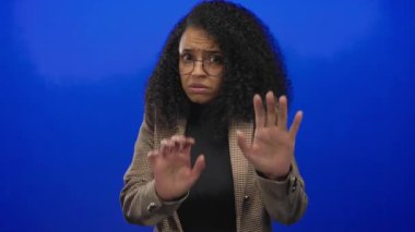 Gözlüklü bir kadın, canlı mavi bir arka plana karşı ellerini kaldırarak reddediyor. İzole edilmiş bir duvarda kıvırcık saçlı ve ekose ceketli bir kadın..