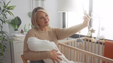 Kadın beşiğin yanında kundaklanmış bebeği tutarken binanın içinde selfie için akıllı telefon tutuyor; annelik bağı.