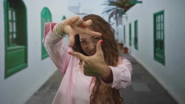 Elleriyle yüzünü çerçeveleyen ve yeşil pencereleri ve saksısı bitkileri olan beyaz duvarlı bir sokakta gülümseyen bir kadın..