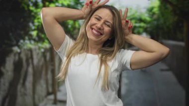 Sarı saçlı bir kadın yeşil yaprakların yanında taş duvarlarla kaplı şehir sokağında elleri ile tavşan kulağı hareketi yapıyor..