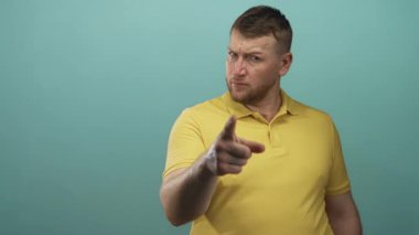 Stüdyoda turkuaz arka plan ve sarı polo tişörtle kamerayı gösteren adam, işaret parmağı izleyiciye doğru uzanıyor; suçlama.