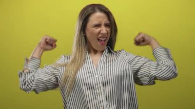 Sarı stüdyoda kaslarını kasarken ve yumruklarını sıkarken gülümseyen kadın çizgili gömlek giyiyor..